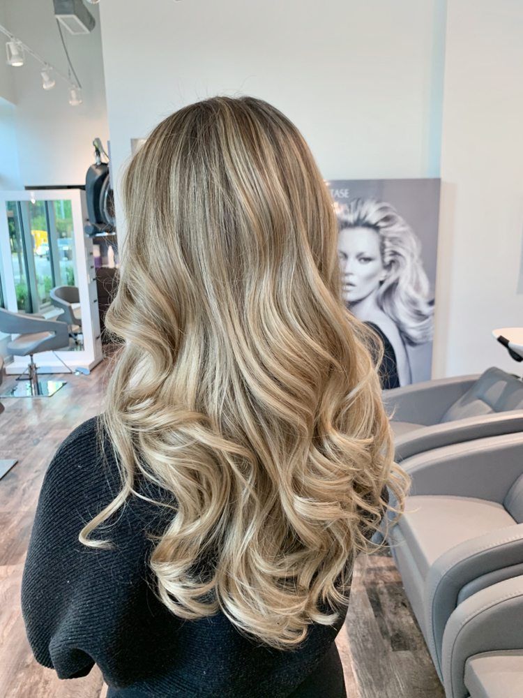 Woman with long, wavy blonde and light brown highlighted hair extensions at a salon.