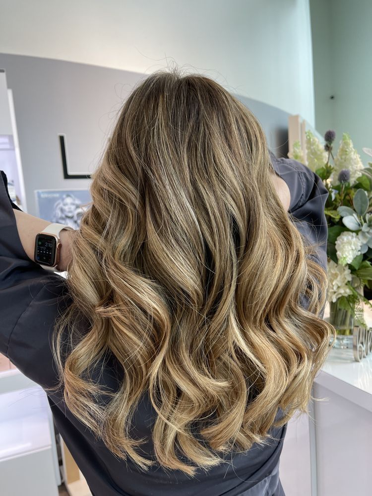 Woman with long, wavy, highlighted brown and blond hair extensions in a salon, hands near the head.
