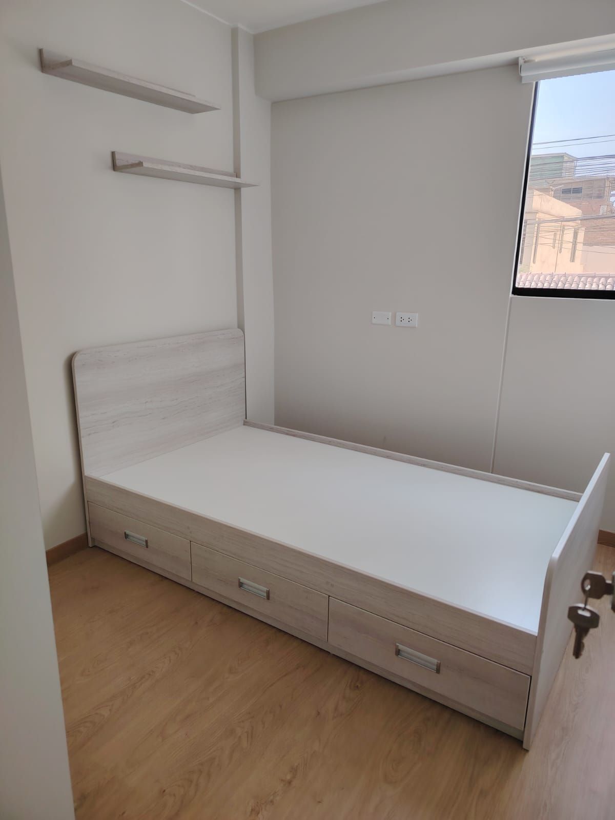 Dormitorio con estructura de cama blanca, cajones y dos estantes en una pared de color claro.