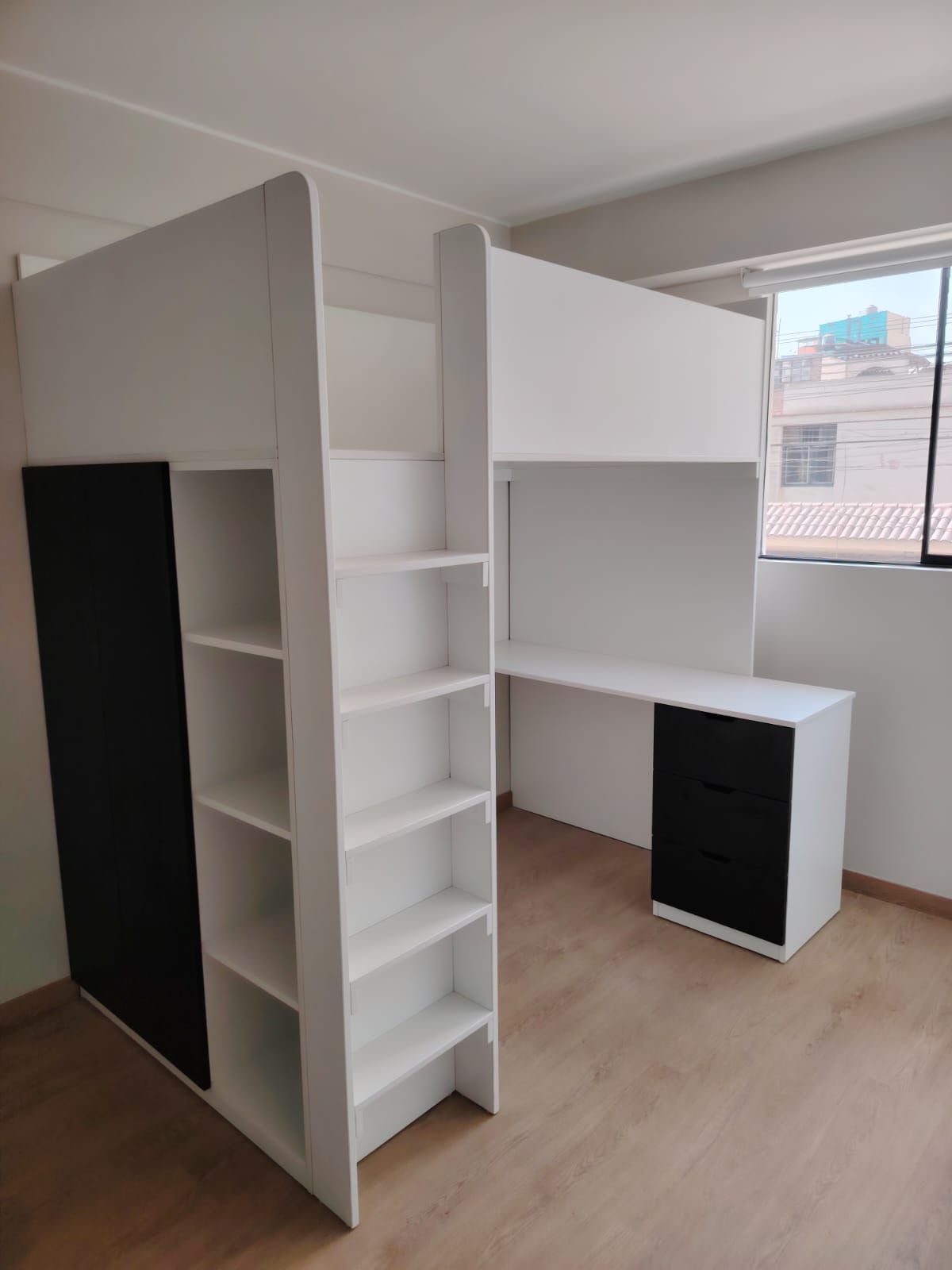 Cama alta blanca con escritorio y estantes de almacenamiento, puerta con detalles negros, piso de madera y una ventana.