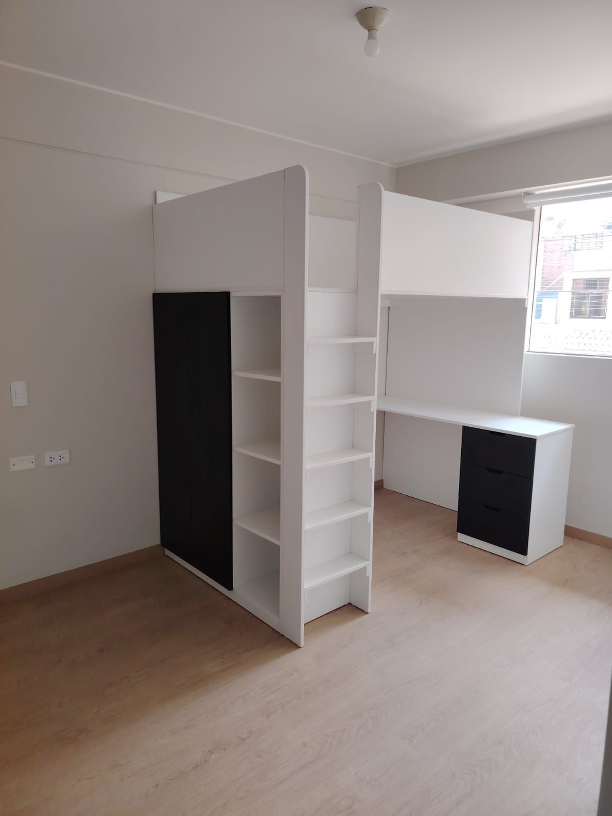 Cama alta blanca con escritorio incorporado, estantes y puerta de armario negra en una habitación con ventana.