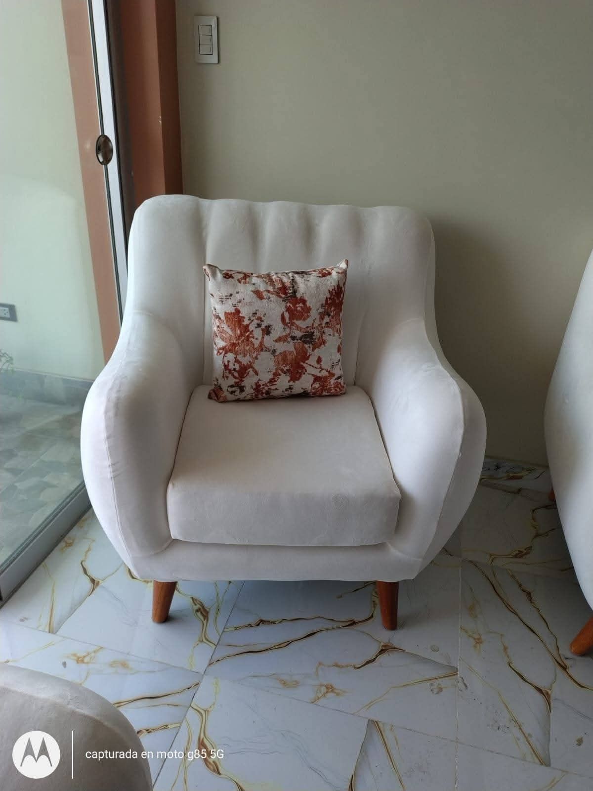 Sillón color crema con cojín floral; patas de madera. Se asienta sobre un suelo de baldosas junto a una puerta de cristal.