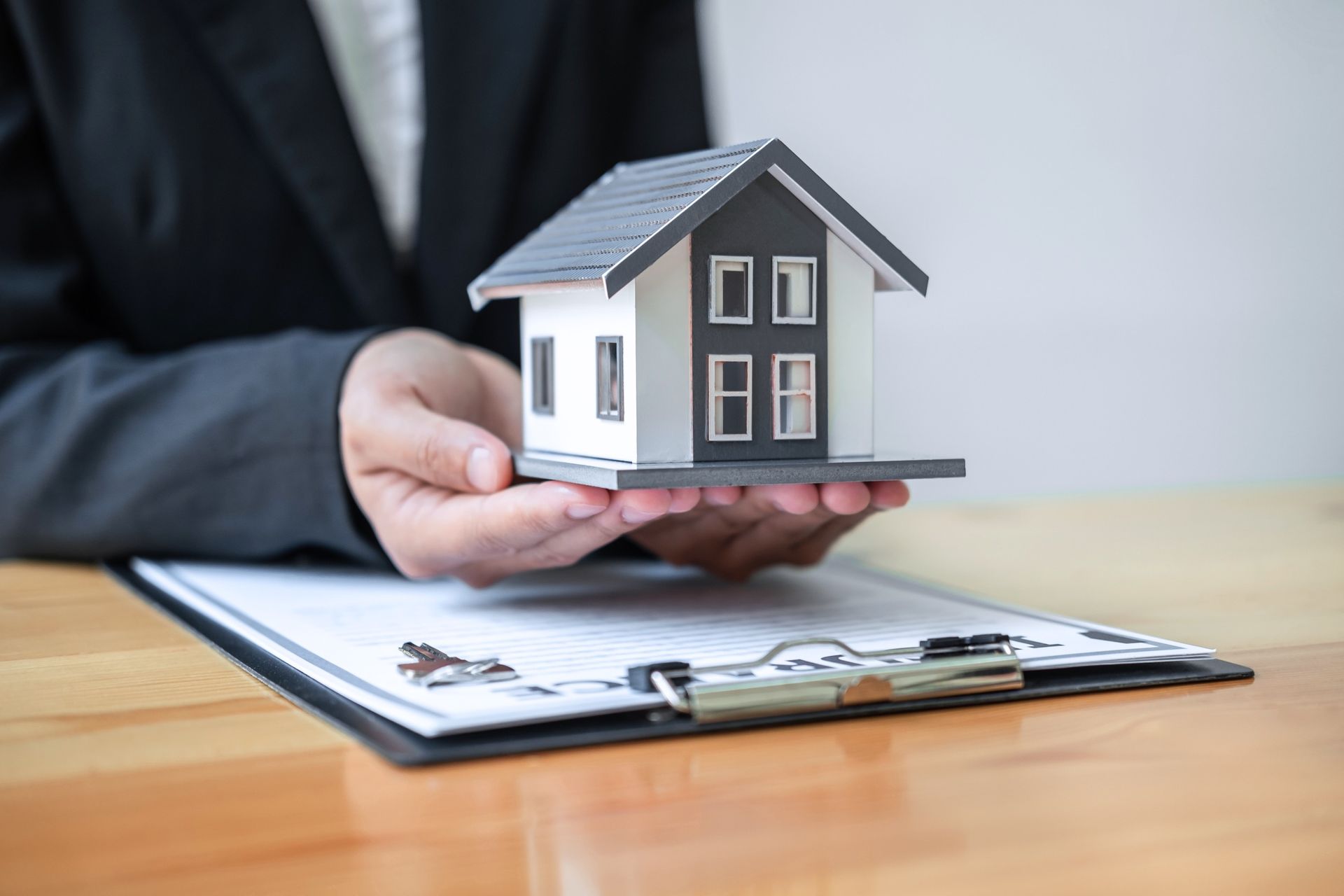 Hand holding a small house model over a clipboard on a desk, suggesting a real estate meeting