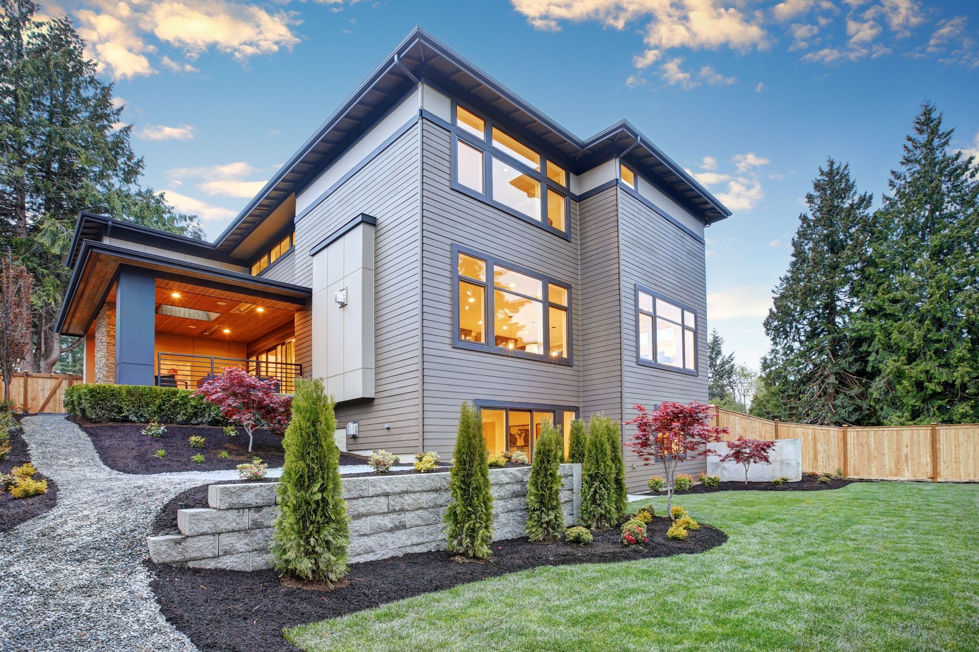 Modern two-story house with gray siding, warm-lit windows, stone steps, and a landscaped front yard at dusk