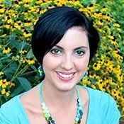 Smiling person with short dark hair in turquoise top, standing in front of yellow flowers