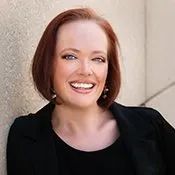Smiling person with short red hair wearing a black top against a light stone wall