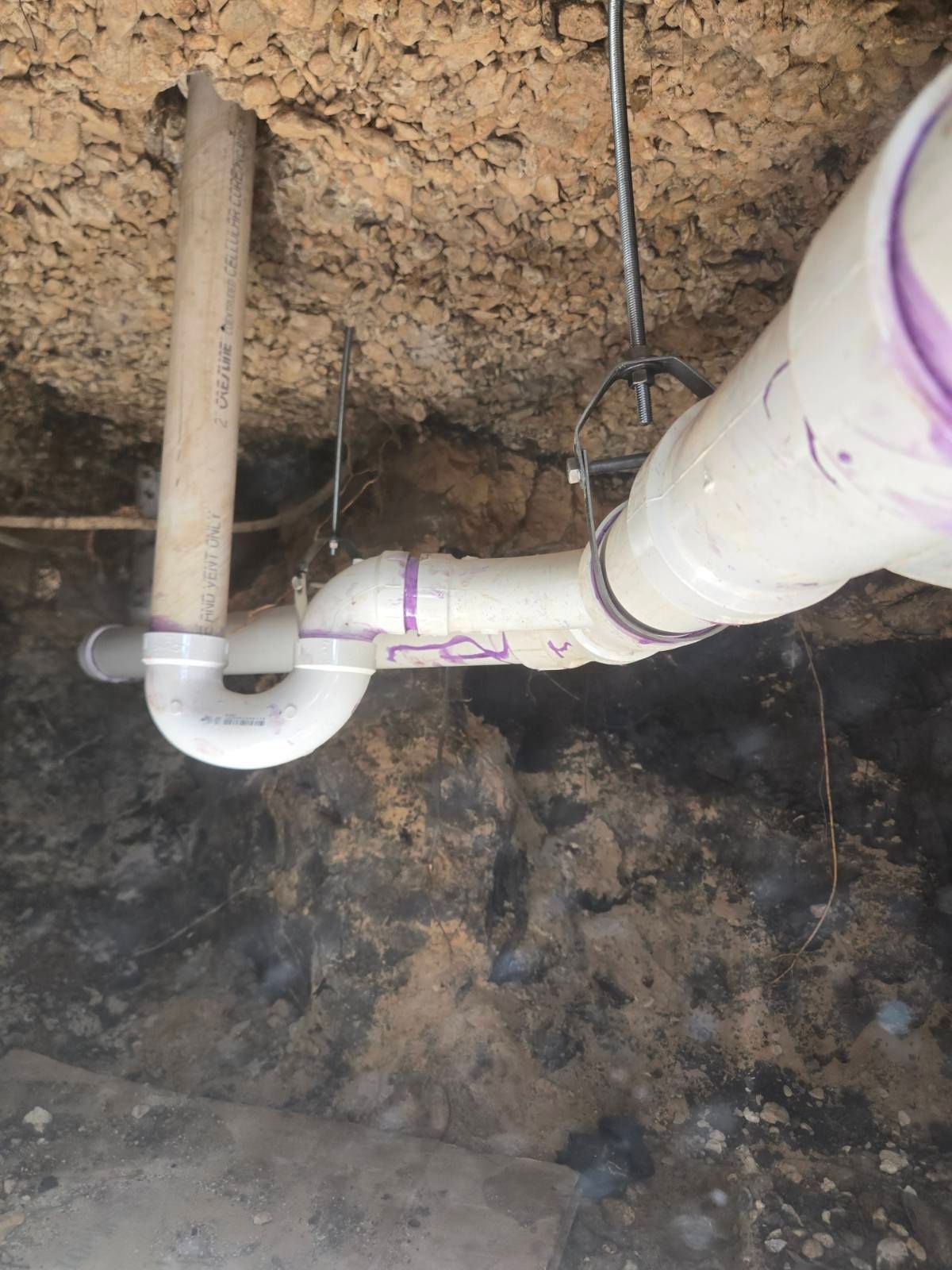 White PVC plumbing pipes and a vertical vent pipe under a dirt crawlspace ceiling.