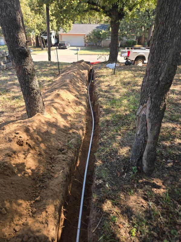 A deep trench runs between two trees in a yard, with a house and parked truck in the background.