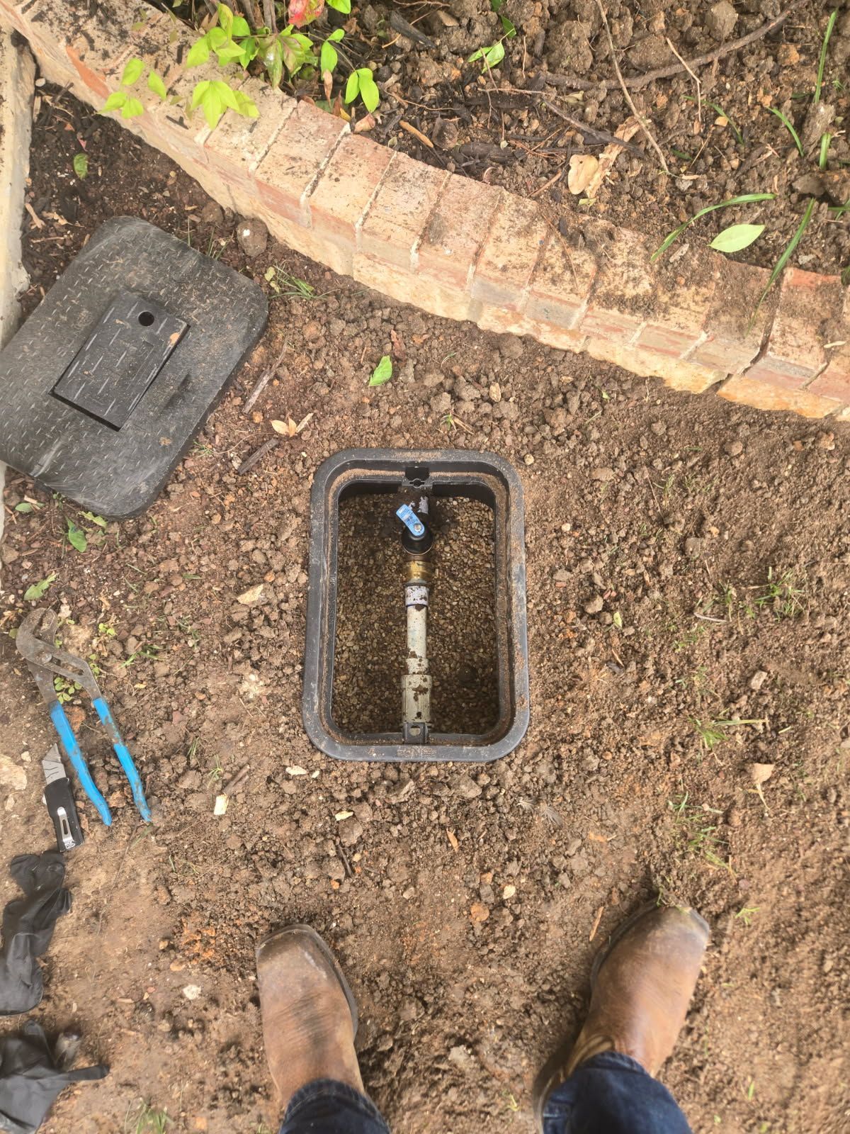 Open utility box in dirt beside curved brick edging, with tools and boots visible nearby.