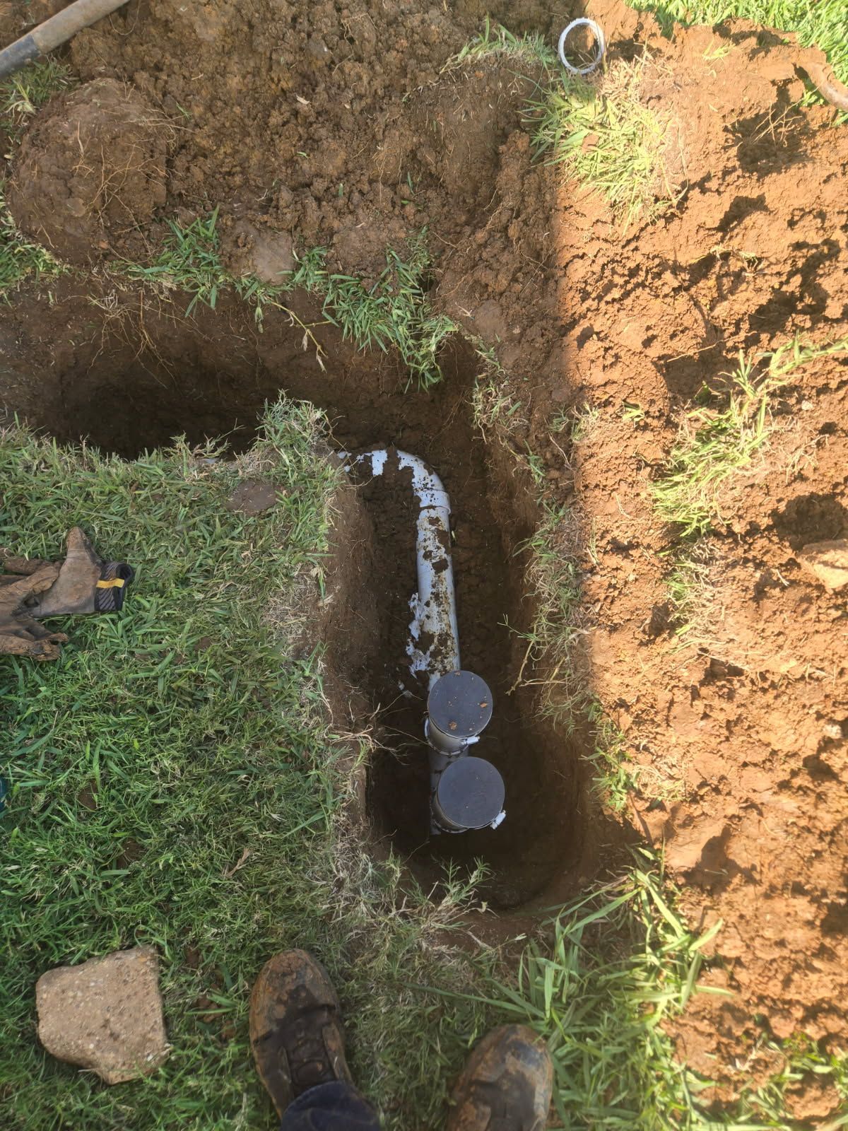 A narrow trench with exposed pipe and water, bordered by grass and soil, with a person's shoes visible at the edge