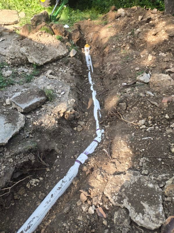 White PVC pipe laid in a dirt trench beside rocks on a sloped outdoor ground.