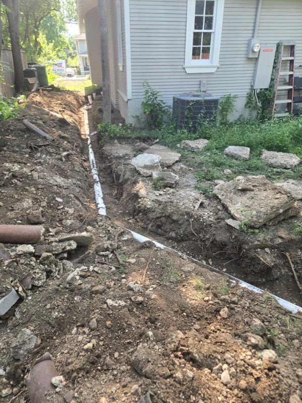 Trenched dirt beside a house foundation with exposed pipe and scattered rocks in a yard