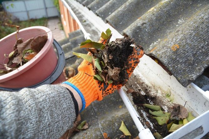 Gloved hand pulling wet leaves and debris from a roof gutter beside a tiled roof