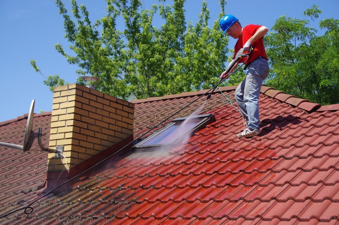Worker pressure-washing a red tiled roof next to a brick chimney on a sunny day