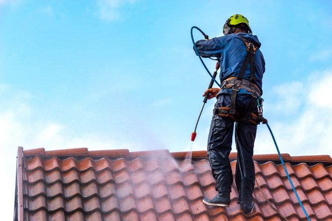 Worker pressure-washing a red-tiled roof while standing on a harness, spraying water into the sky.