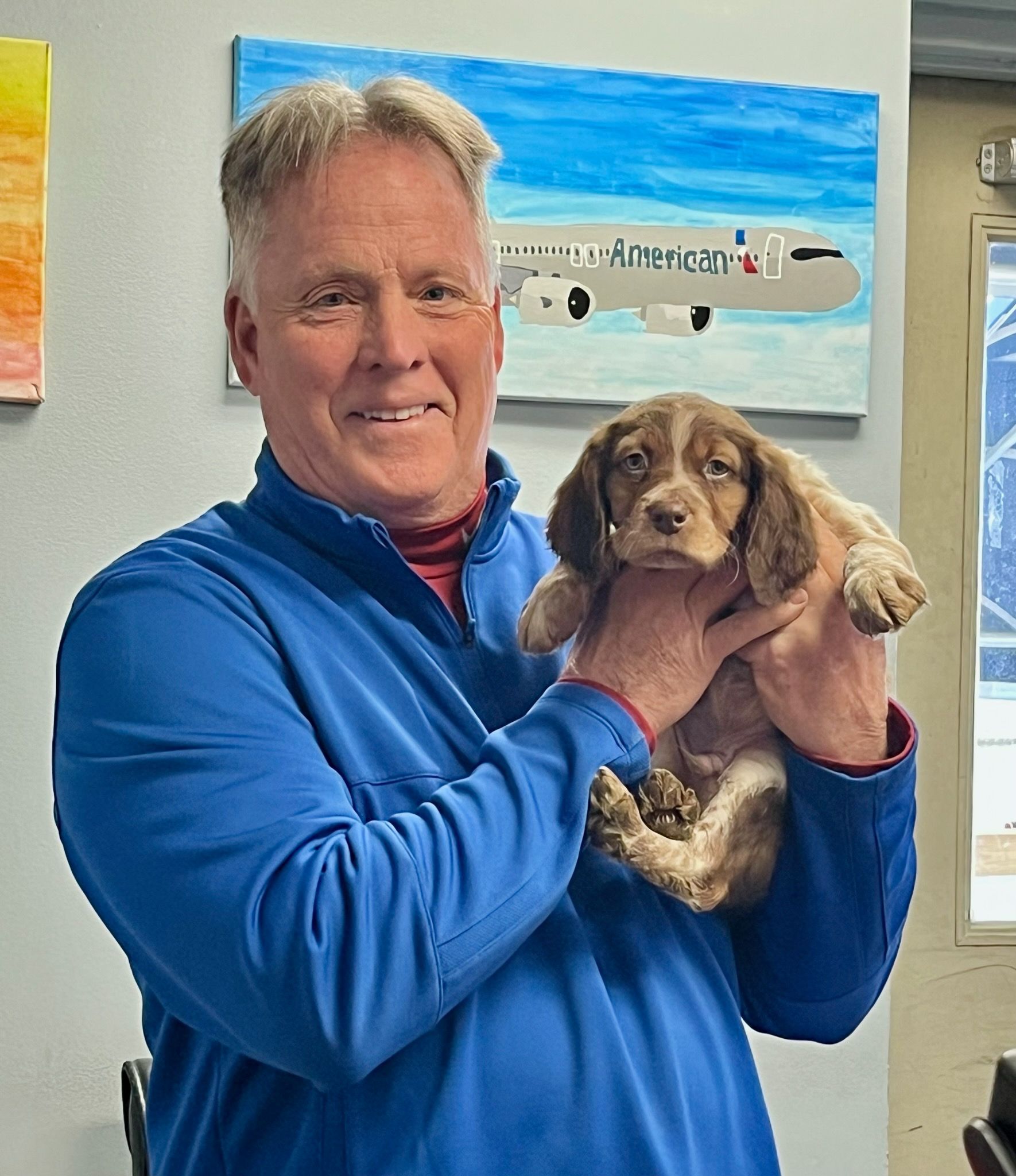 Man in blue holding a puppy with brown and white fur, smiling indoors with paintings visible.