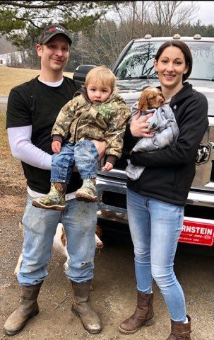 Family standing in front of a truck; man holding a child in camo jacket, woman holding a dog.