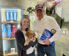 Woman and man holding puppy and book, standing by ribbons and a turkey mount.