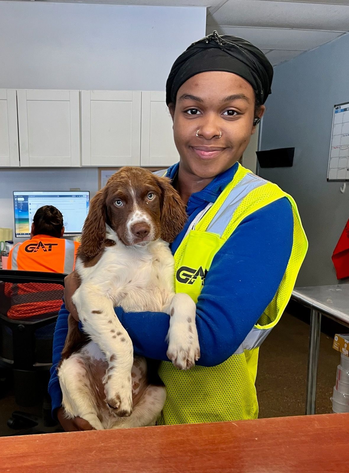 Woman in blue and yellow vest holds a brown and white puppy; office setting with computer in the background.
