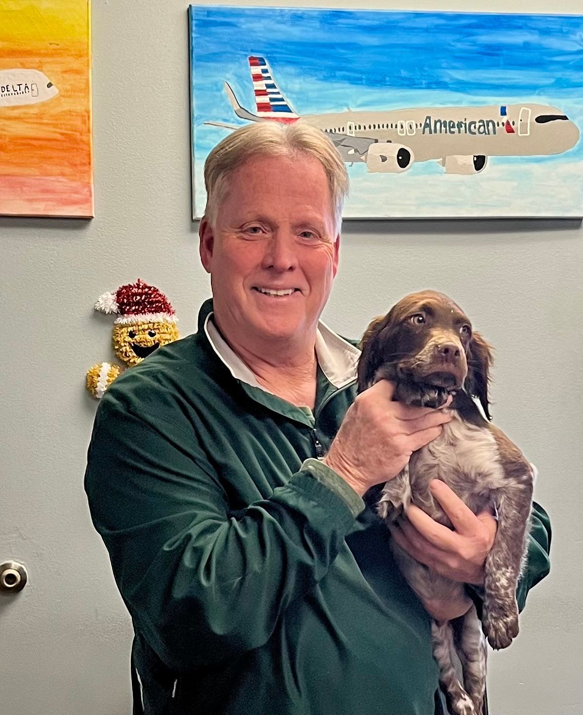 Man in green shirt smiles, holding a brown and white puppy. Paintings hang on the wall.