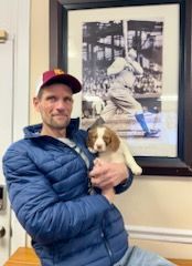 Man in blue jacket holding puppy, posing in front of a framed vintage baseball photo.