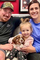 Family of four, including a young child holding a puppy, seated on a couch; all smiling.