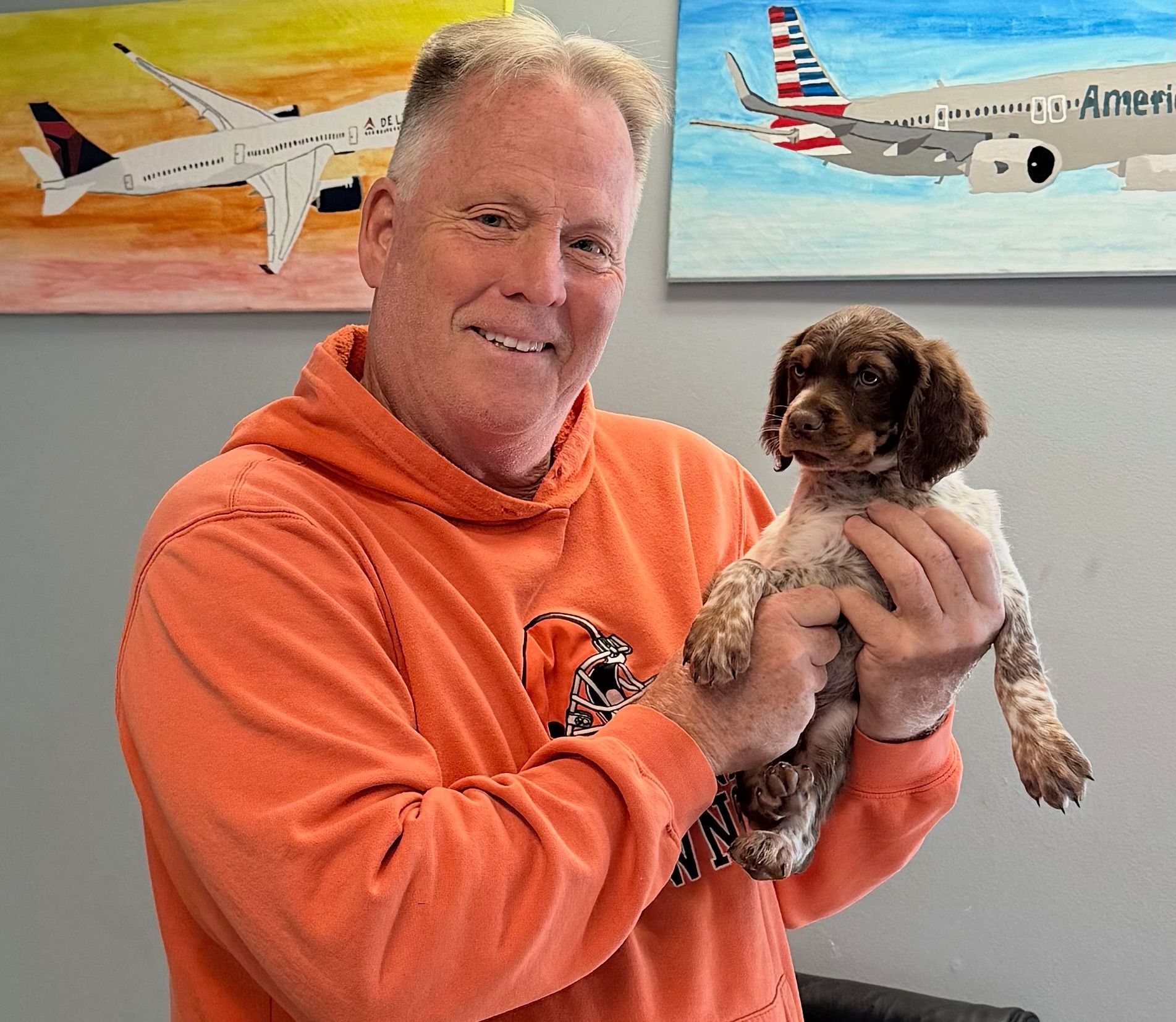 Man in orange hoodie holding a small brown and white dog; two airplane paintings in the background.
