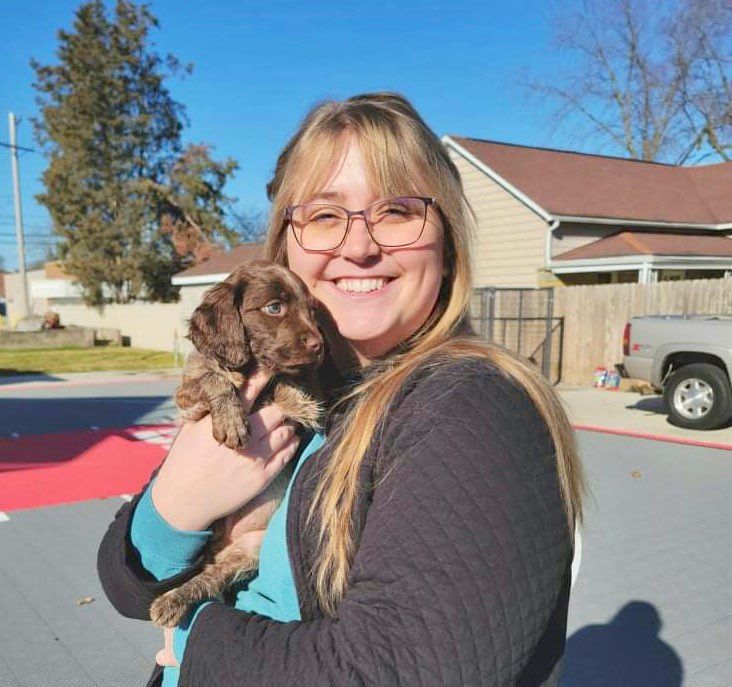 Woman smiling, holding a brown puppy outdoors near a building; sunny day.