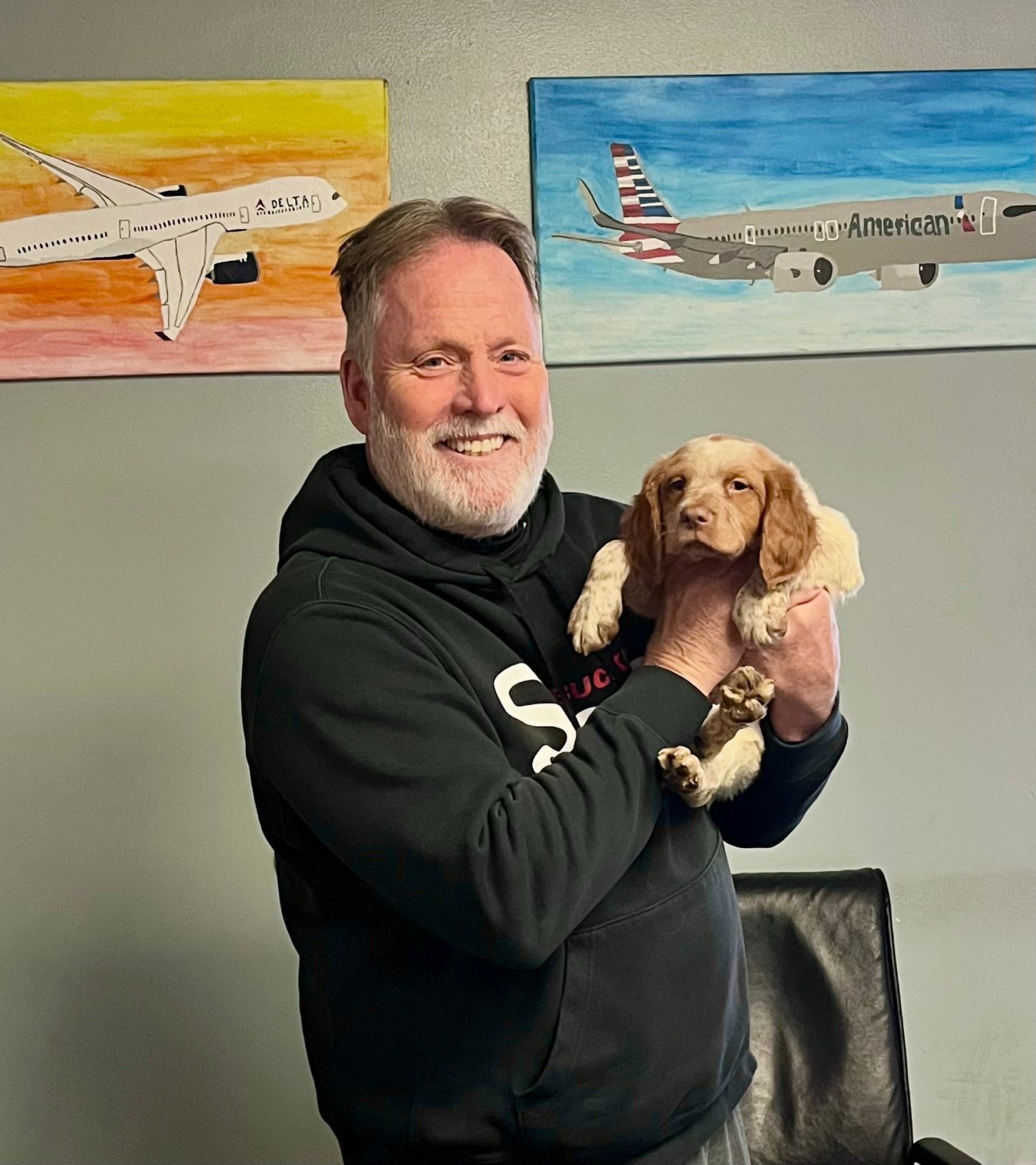 Man holding a puppy in front of airplane paintings, smiling.
