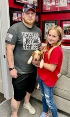 Man holding a puppy with a girl in a red shirt. Background has framed items.