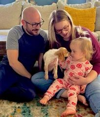 Family sitting with a puppy and child. Parents are smiling and looking at the dog and child.