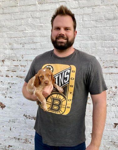 Man holding a small, brown and white puppy. Both smile in front of a white brick wall. Man wears a Bruins t-shirt.