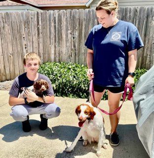 Two people with dogs outdoors; one person holds a puppy, the other holds the leash of a sitting dog.