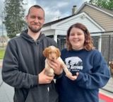 Couple holding a small, brown and white puppy outdoors, smiling.