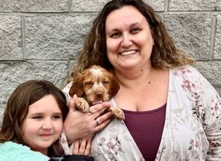 Woman and young girl smiling with a puppy. The woman holds the puppy outdoors.