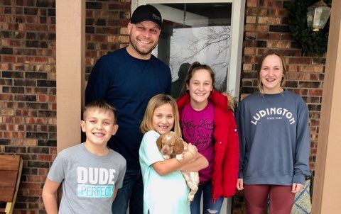 Family of five and a puppy pose in front of a house. The girl holds the dog.