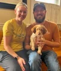 Couple seated, holding a puppy. Woman smiles in a yellow shirt, man in an orange shirt. Indoors, natural light.