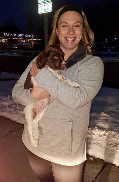 Woman holding a brown and white puppy. Smiling outdoors with snow and a restaurant sign.