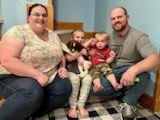 Family of four sitting together. Woman in floral top, man with beard, two young children, indoors.