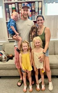 Family of five with a puppy poses near a decorative wall. Two young girls and a toddler are in the photo.