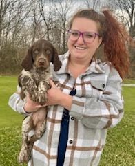 Woman with glasses smiling, holding a speckled puppy outdoors.