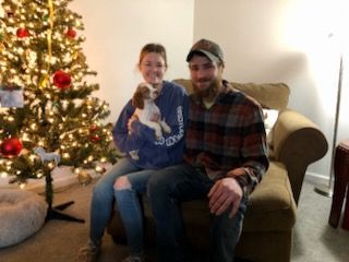 Couple seated on a couch with a small dog, in front of a Christmas tree with ornaments.