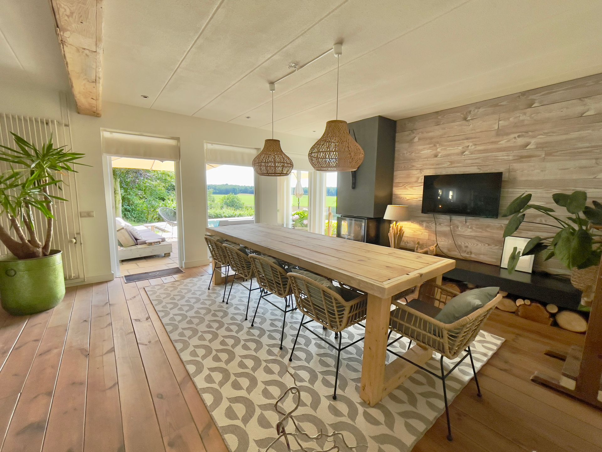 Dining room with long wooden table, wicker chairs, and woven pendant lights. Fireplace and open door with outdoor view.