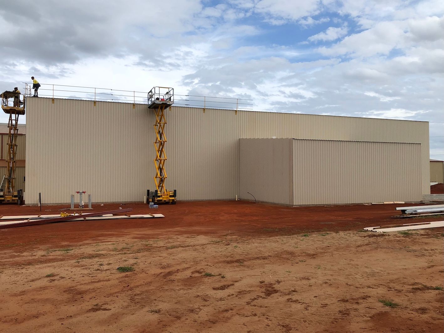 Large beige industrial building; workers on lifts, installing roof components. Cloudy sky, dirt ground.