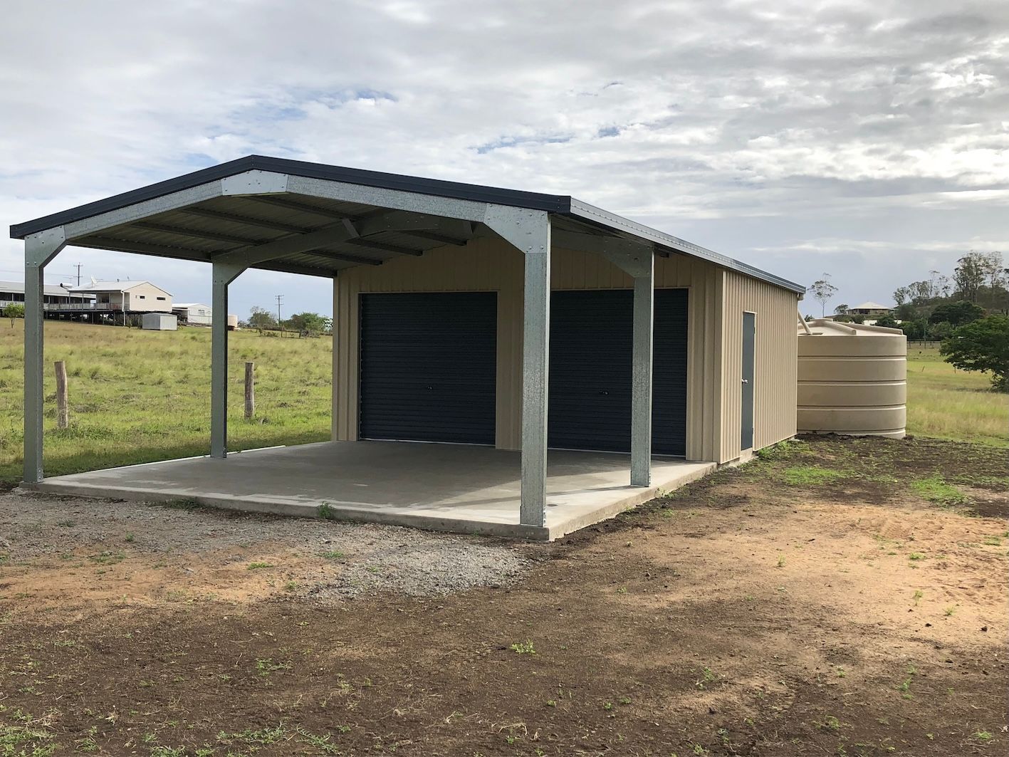 A tan metal shed with a carport. It has two roller doors, and a water tank, on a concrete pad.