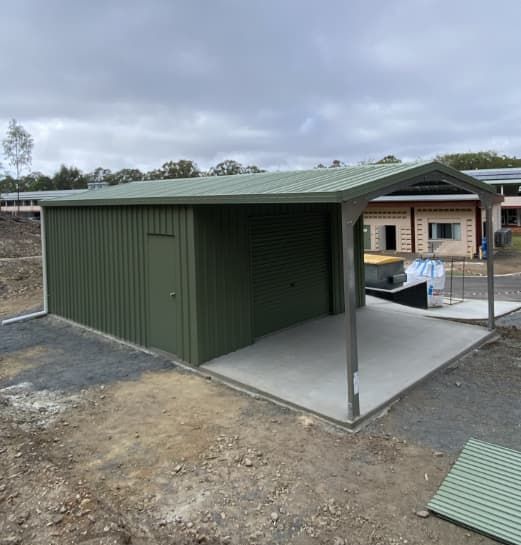 A Green Shed With A Roof Is Sitting In The Dirt In Front Of A House — Shed Constructions (QLD) In Gin Gin, QLD