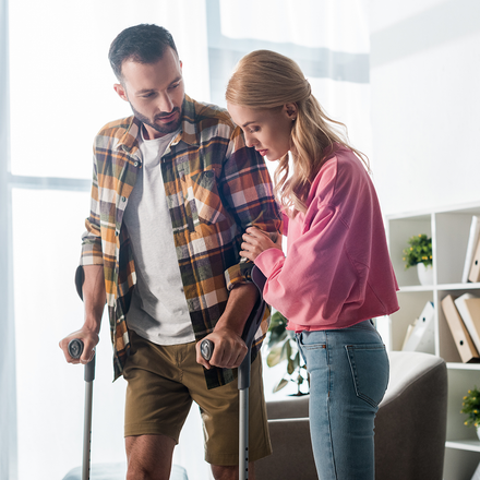 A woman is helping a man with crutches walk in a living room.