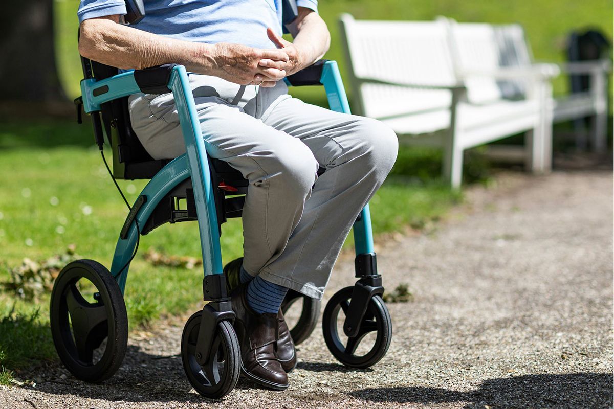 Man seated in a blue rollator walker on a paved path outdoors, hands clasped.