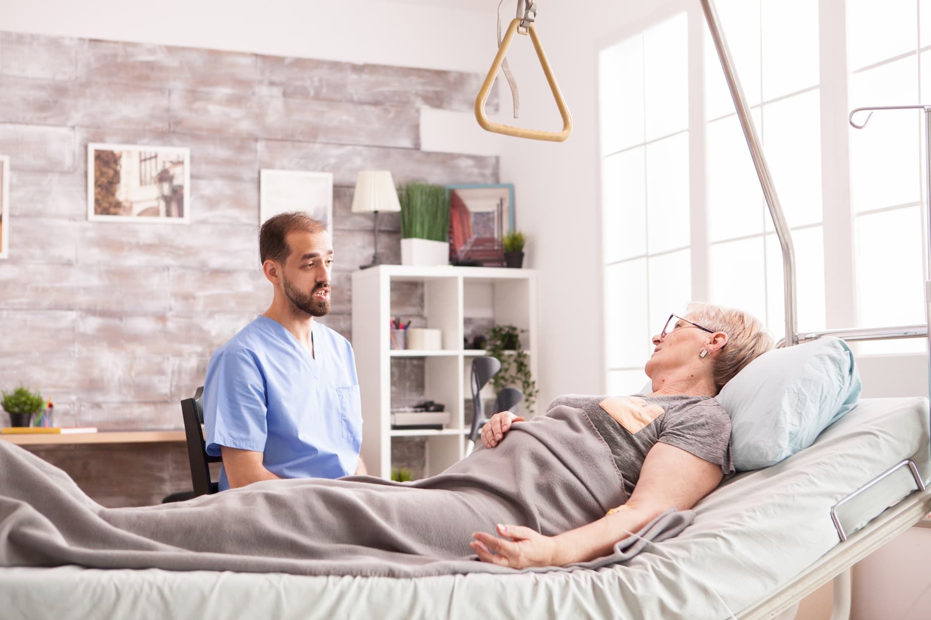 An occupational therapist is talking to an elderly woman laying in a hospital bed.