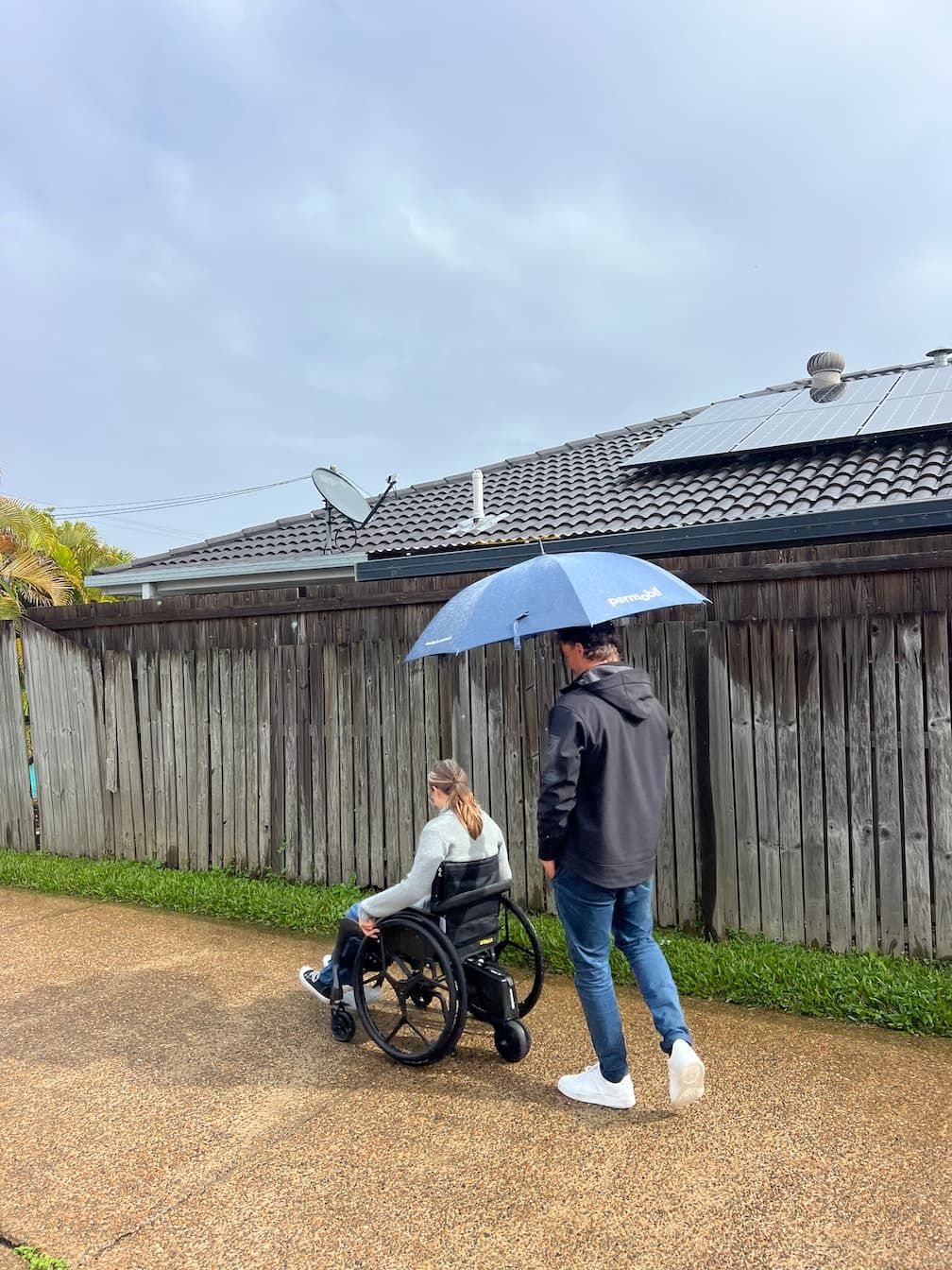 An occupational therapist is walking a woman in a wheelchair under an umbrella.